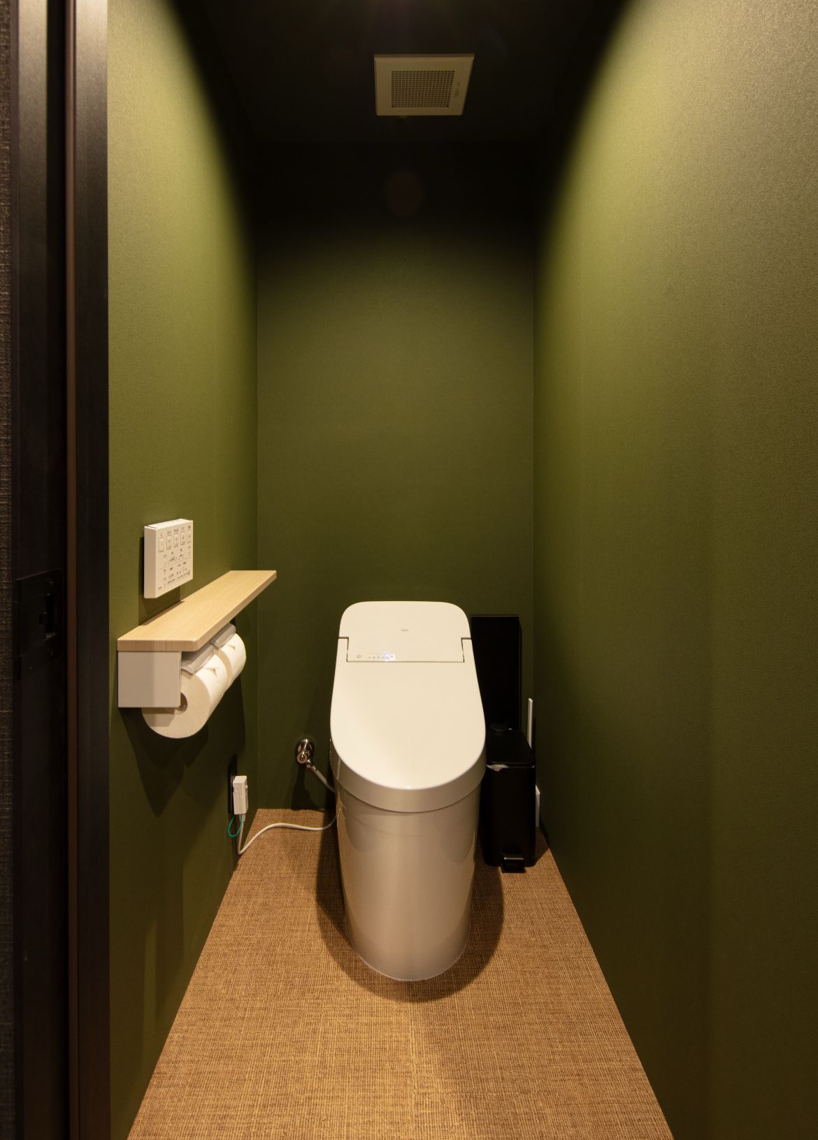 A small, narrow bathroom with a toilet, a paper towel dispenser, and a trash bin.