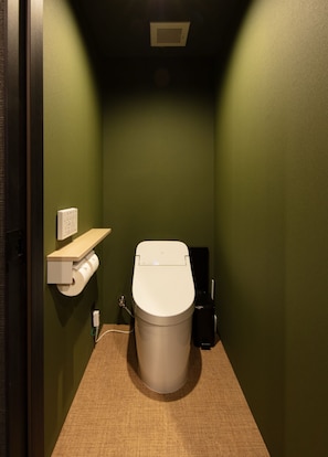 A small, narrow bathroom with a toilet, a paper towel dispenser, and a trash bin.