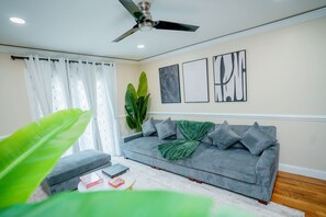 A living room with a grey sofa, a ceiling fan, and a large green plant. - Modern Luxury 3BR | 2 Baths | Parking | Near NYC (Jersey City)