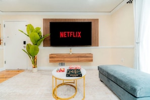 A living room with a television displaying 'NETFLIX', a small round table with chess and board games, a green plant, and a blue sofa. - Modern Luxury 3BR | 2 Baths | Parking | Near NYC (Jersey City)