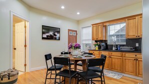A dining area with a round table, four chairs, a vase with flowers, and a kitchen with wooden cabinets and a window.