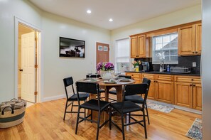 A dining area with a round table, four chairs, a vase with flowers, and a kitchen with wooden cabinets and a window.