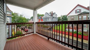 A wooden porch with a railing, overlooking a residential street with houses and parked cars.