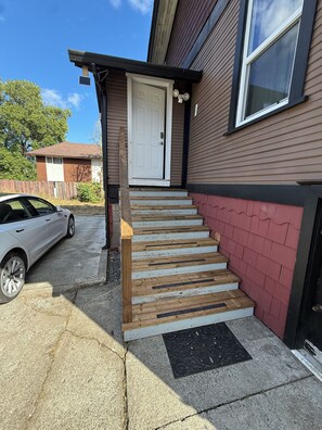 Un immeuble résidentiel avec une porte d’entrée, un escalier en bois menant à l’entrée et une voiture garée dans l’allée.