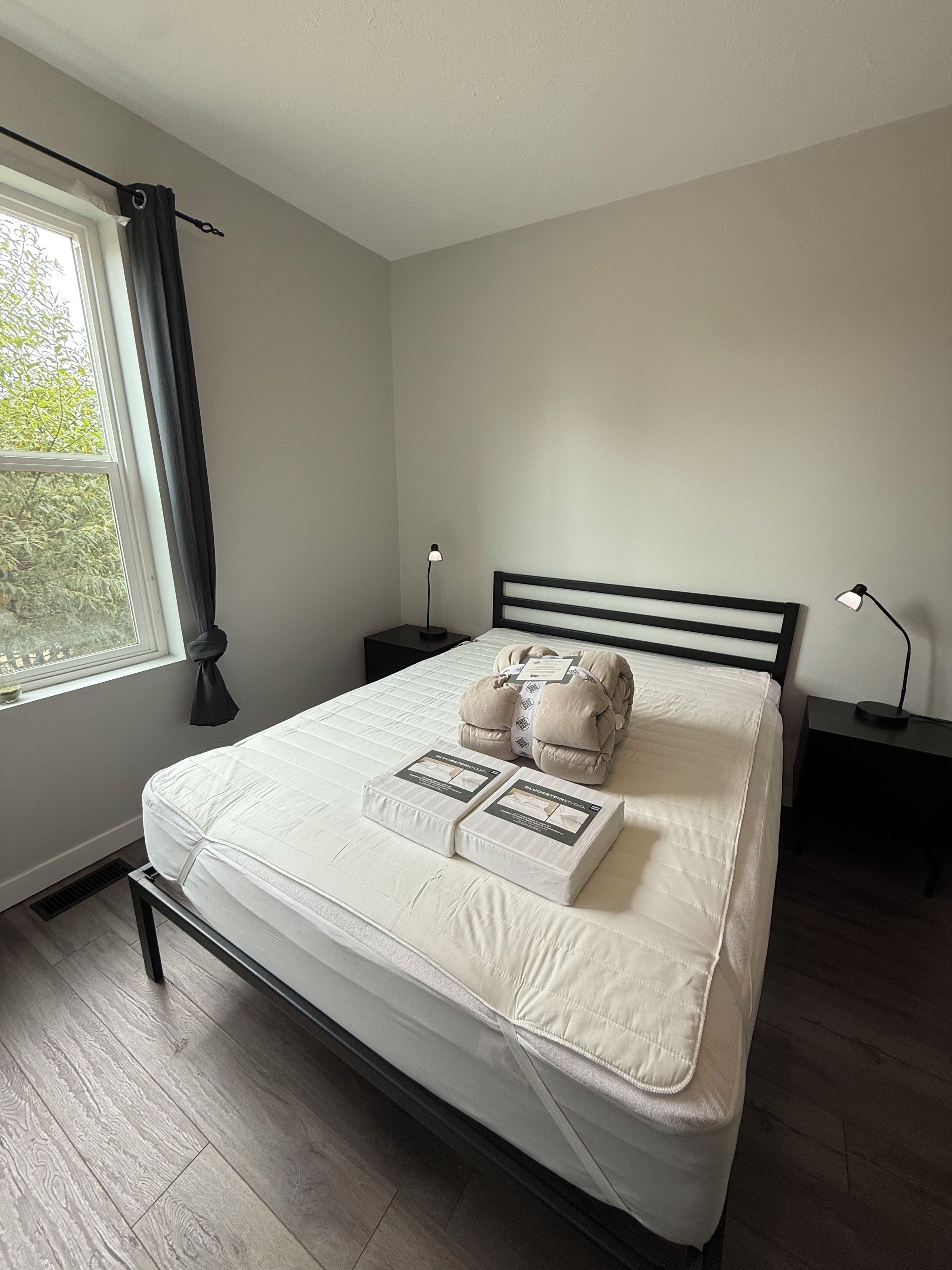 A neatly made bed with pillows and a blanket, a bedside table with a lamp, a window with curtains, and a view of greenery outside.