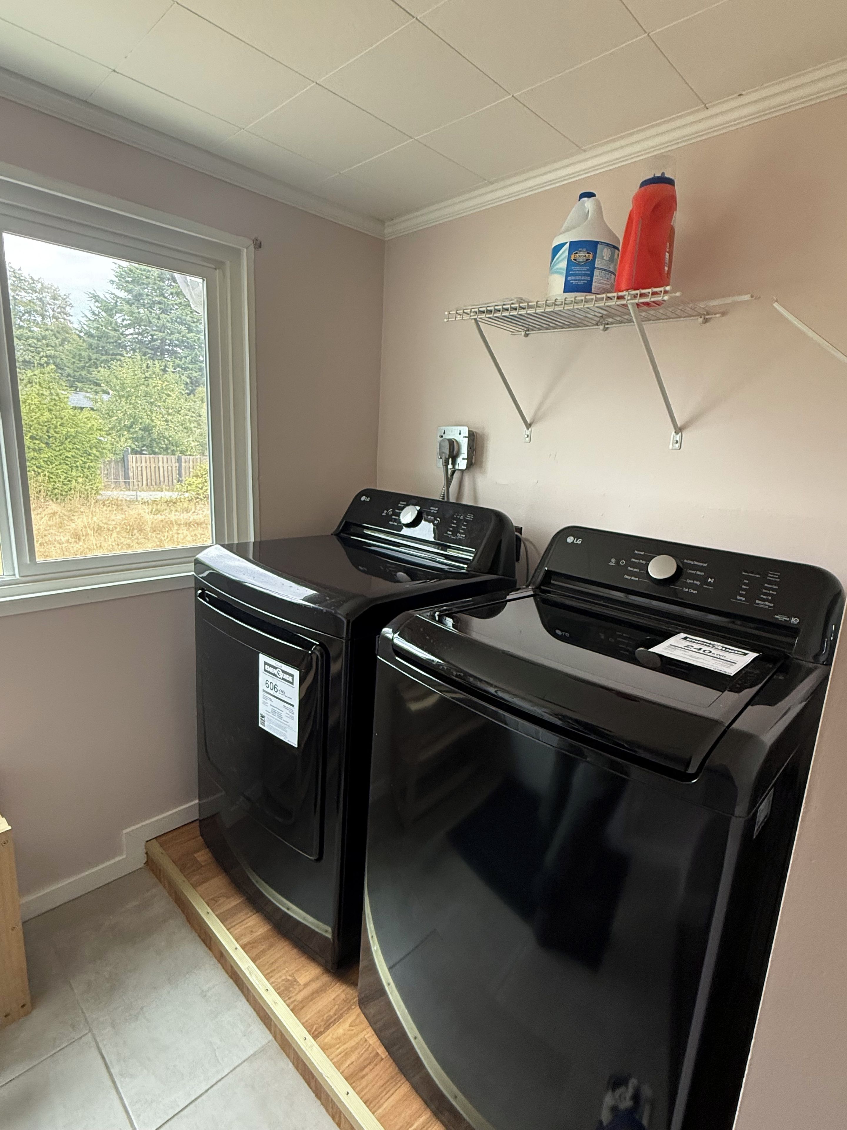 A laundry room with two black washing machines, a window, and a shelf with cleaning supplies.
