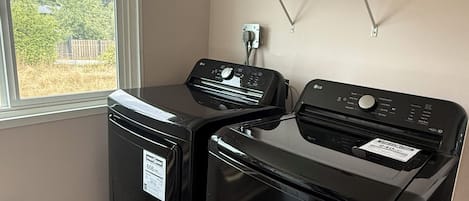 A laundry room with two black washing machines, a window, and a shelf with cleaning supplies.