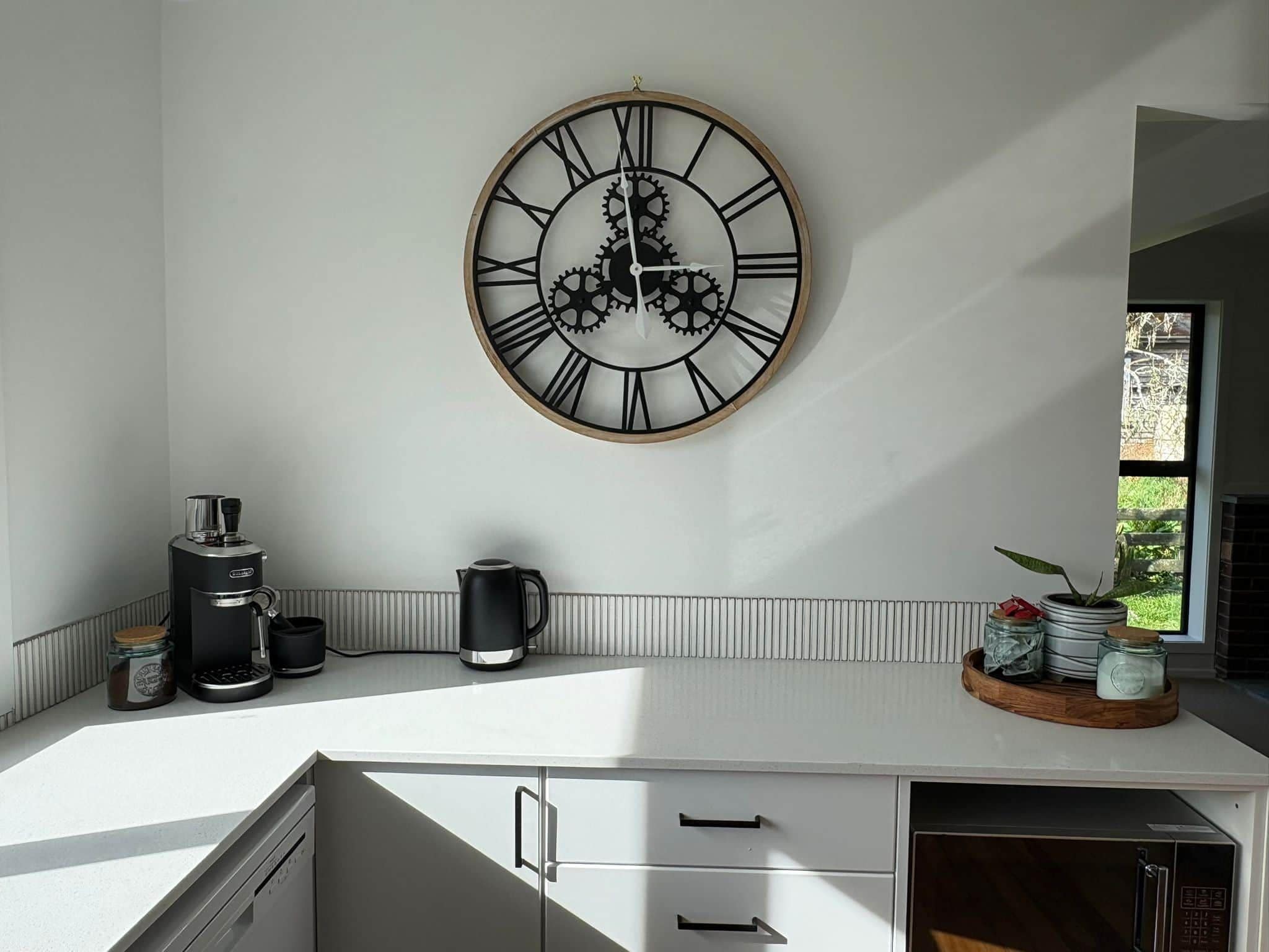 Una cocina con un reloj en la pared, una cafetera, un hervidor y una ventana con vistas a zonas verdes.