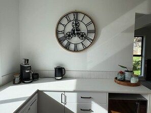 Une cuisine avec une horloge murale, une machine à café, une bouilloire et une fenêtre donnant sur un paysage verdoyant.