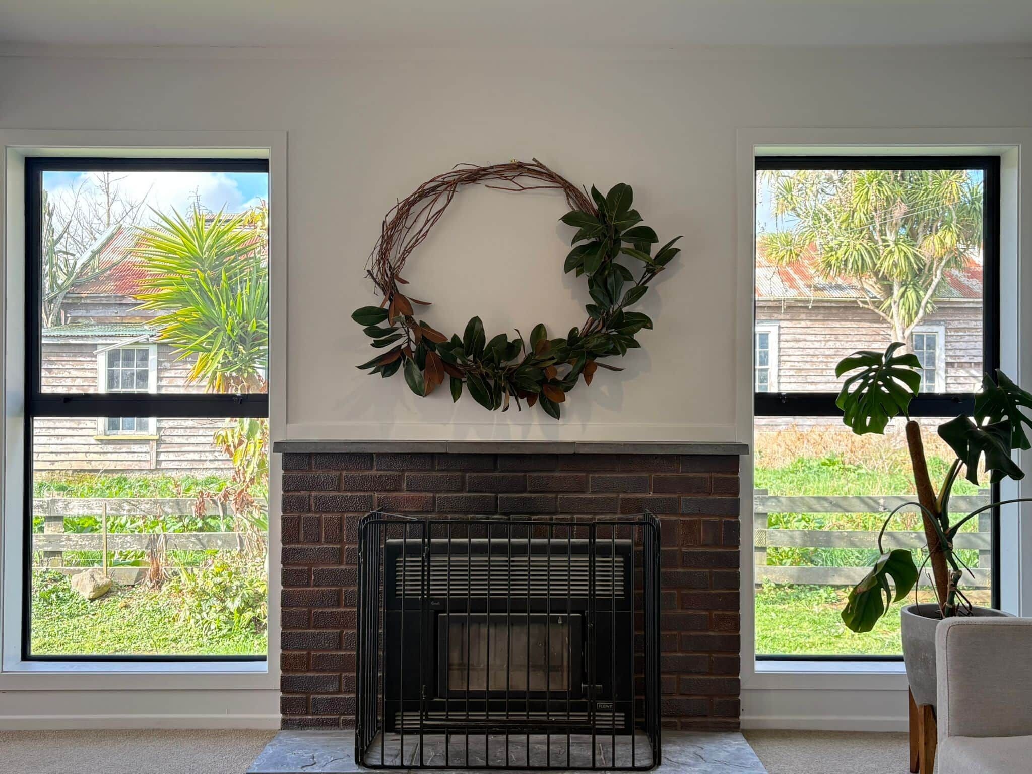 A room with a fireplace, two windows with views of a garden and a house, and a wreath on the wall.