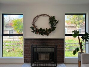Una habitación con chimenea, dos ventanas con vistas a un jardín y una casa, y un adorno en la pared.