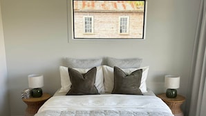 Una cama bien hecha con ropa blanca y dos almohadas, flanqueada por dos mesitas de noche con lámparas, en una habitación con una pared lisa y una ventana con vistas a un edificio.