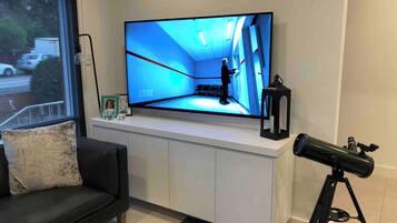 Una sala de estar moderna con un televisor de pantalla plana montado en un mueble blanco, un telescopio sobre un trípode y un sofá negro con un cojín.