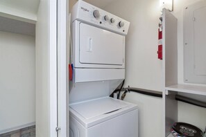 A compact laundry room with a stacked washer and dryer, a fire extinguisher, and a metal shelf. - Ocean Front w/Direct Pvt Access, Free Beach Setup! New Owner, Pre-Reno Special! (Destin)