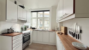 Eine kleine Küche mit weißen Schränken, einem eingebauten Backofen, einem Fenster mit Blick auf Gebäude und einem Messerblock an der Wand.