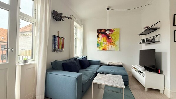 Ein modernes Wohnzimmer mit einem Ecksofa, einem kleinen Couchtisch, einem Flachbildfernseher und einer an der Wand hängenden Gitarre.