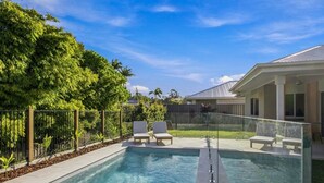 En swimmingpool med en glasafskærmning, omgivet af et hegn, og beliggende ved siden af en bygning med en overdækket terrasse.