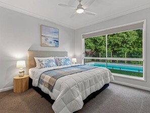 A bedroom with a bed, a ceiling fan, a window with a view of a swimming pool and greenery.