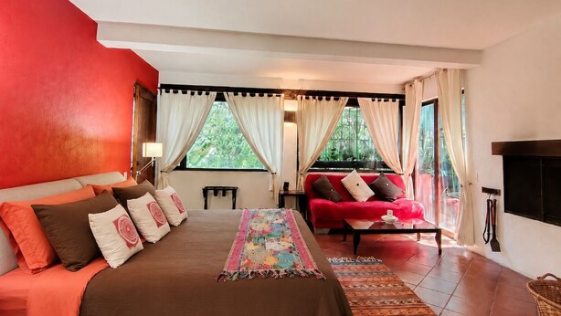 A bedroom with a large bed, a red sofa, and a view of greenery through the windows. - Quetzal Suite | Terrace, fireplace and jacuzzi (Valle de Bravo)