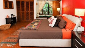 A bedroom with a bed, pillows, a nightstand, a lamp, a window with curtains, a wooden wardrobe, and a small table with a basket.