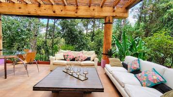 A covered outdoor seating area with a wooden pergola, a white sofa with colorful cushions, a wooden coffee table with candles, and a chair.