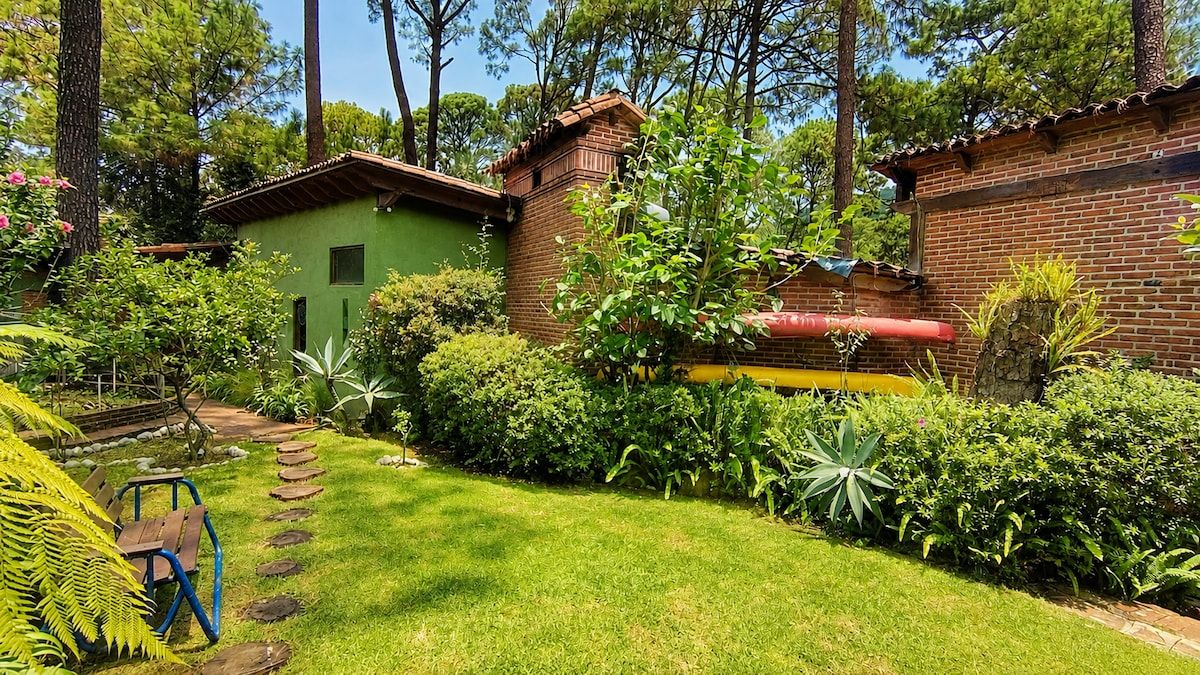 A green house with a brick chimney, surrounded by a well-maintained garden with a variety of plants and a wooden bench.