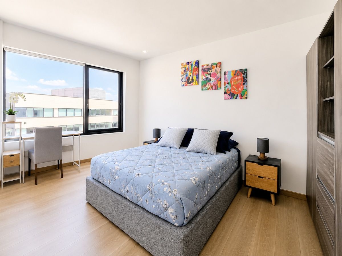 Ein modernes Schlafzimmer mit einem großen Bett, einem Schreibtisch und einem Stuhl. An der Wand hängen drei Bilder und ein Fenster bietet einen Blick ins Freie.