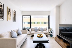 Ett modernt vardagsrum med en soffa, ett soffbord och en tv. På väggen hänger inramade konstverk och det finns ett stort fönster med utsikt över byggnader.