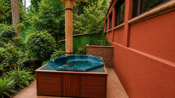 A hot tub in a garden area with a wooden deck, surrounded by lush greenery and a red wall.
