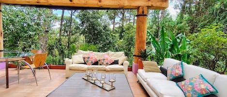 A covered outdoor seating area with a wooden pergola, a white sofa with colorful cushions, a wooden coffee table with candles, and a chair.