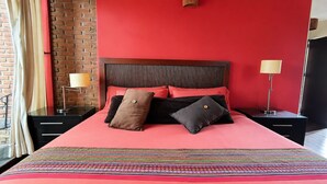 A hotel room with a red wall, a wooden headboard, a bed with a colorful blanket, two bedside tables with lamps, and a brick wall. - Paloma Suite | Triple, breakfast and parking (Valle de Bravo)