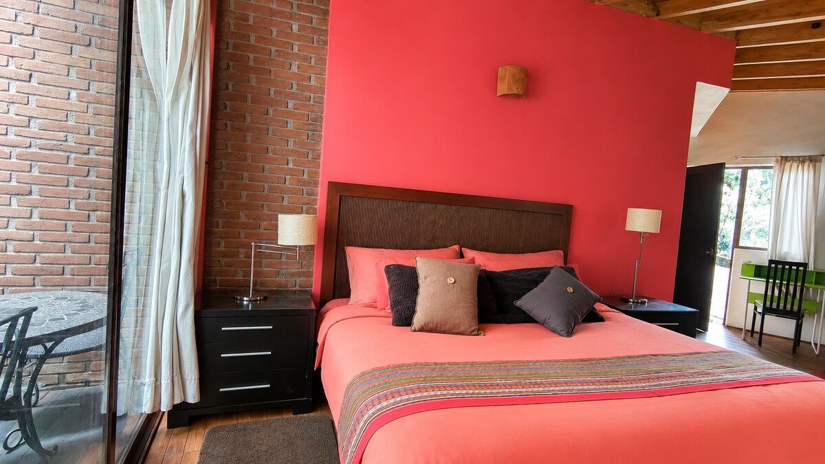 A bedroom with a brick wall, a large bed with multiple pillows, a nightstand with a lamp, and a view of the outdoors.