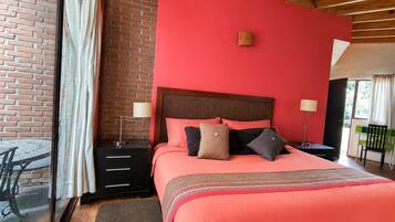 A bedroom with a brick wall, a large bed with multiple pillows, a nightstand with a lamp, and a view of the outdoors.