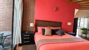 A bedroom with a brick wall, a large bed with multiple pillows, a nightstand with a lamp, and a view of the outdoors.