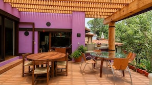 An outdoor patio with a round wooden table and chairs, surrounded by potted plants and a pergola structure.