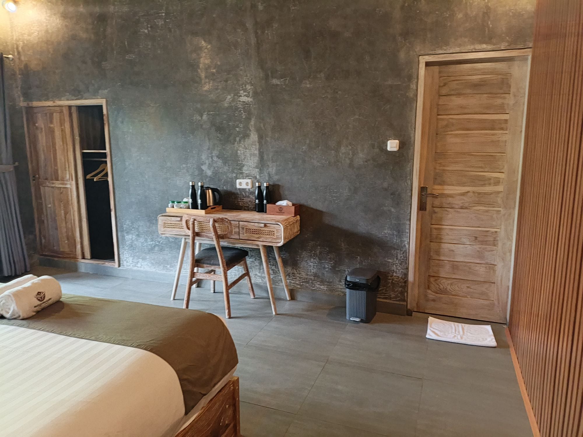 Une pièce comprenant un lit, un bureau en bois avec des bouteilles et une bouilloire, une poubelle et une porte.