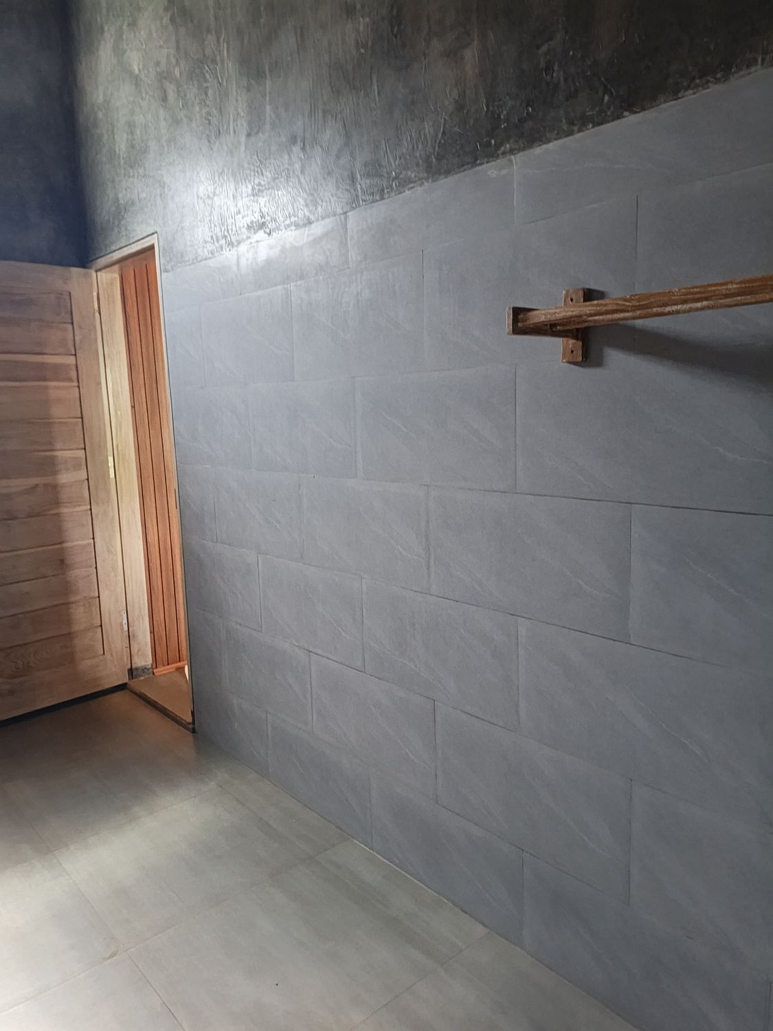 A room with grey tiled walls, a wooden door, and a wooden beam on the wall.