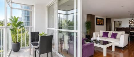 Un vaste espace de vie agrémenté d’un canapé blanc moderne, d’une table à manger et d’une grande porte-fenêtre coulissante donnant sur un balcon où sont disposées des plantes en pot.