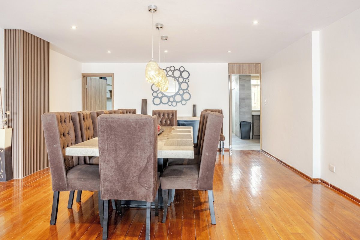 Een moderne eethoek met een houten vloer, een centrale tafel omringd door fauteuils en een decoratieve lamp erboven.