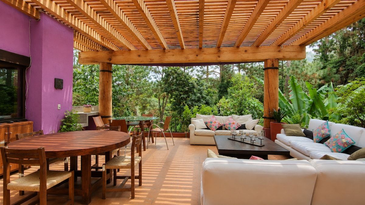 An outdoor patio with a wooden pergola, a round wooden table with chairs, a white sofa with cushions, and a small coffee table.