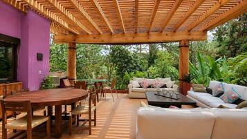 An outdoor patio with a wooden pergola, a round wooden table with chairs, a white sofa with cushions, and a small coffee table.