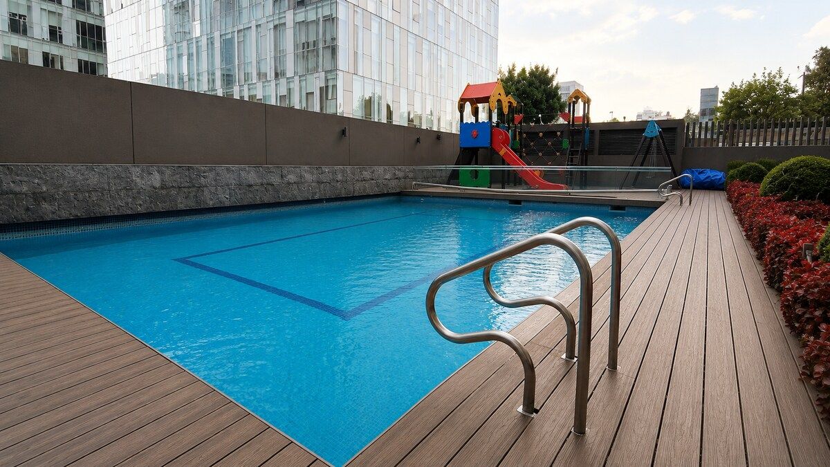 En swimmingpool med en træterrasse, et legeområde for børn og en moderne bygning i baggrunden.
