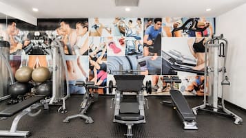 Academia com equipamentos de ginástica, uma parede com fotos relacionadas a exercícios físicos e piso de tapete preto.