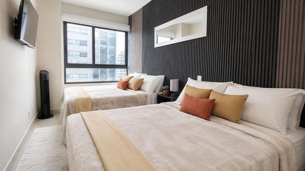 A modern hotel room with two beds, a large window, a flat-screen TV, and a wall-mounted mirror.