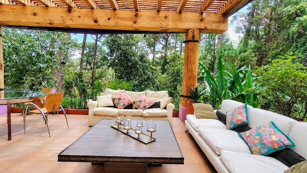 A covered outdoor seating area with a wooden pergola, a white sofa with colorful cushions, a wooden coffee table with candles, and a chair. - Sparrow | King and Breakfast in the Woods (Valle de Bravo)