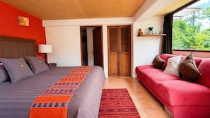 A bedroom with a bed, a red sofa, a wooden wardrobe, and a window with a view of greenery.