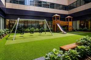 Ein Spielplatz mit Schaukeln und Rutsche, umgeben von Rasenmatte und Kübelpflanzen.