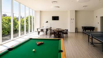 Mesa de bilhar com bolas, mesa de sinuca e mesa de pingue-pongue em um espaço com janelas amplas e vista para o verde.