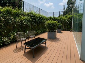 Una terrazza esterna con pavimento in legno, due sedie in vimini, una panca nera e piante in vaso.