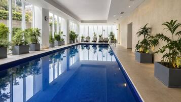 Uma piscina interior extensa com paredes de vidro transparente, plantas em vasos e design moderno.